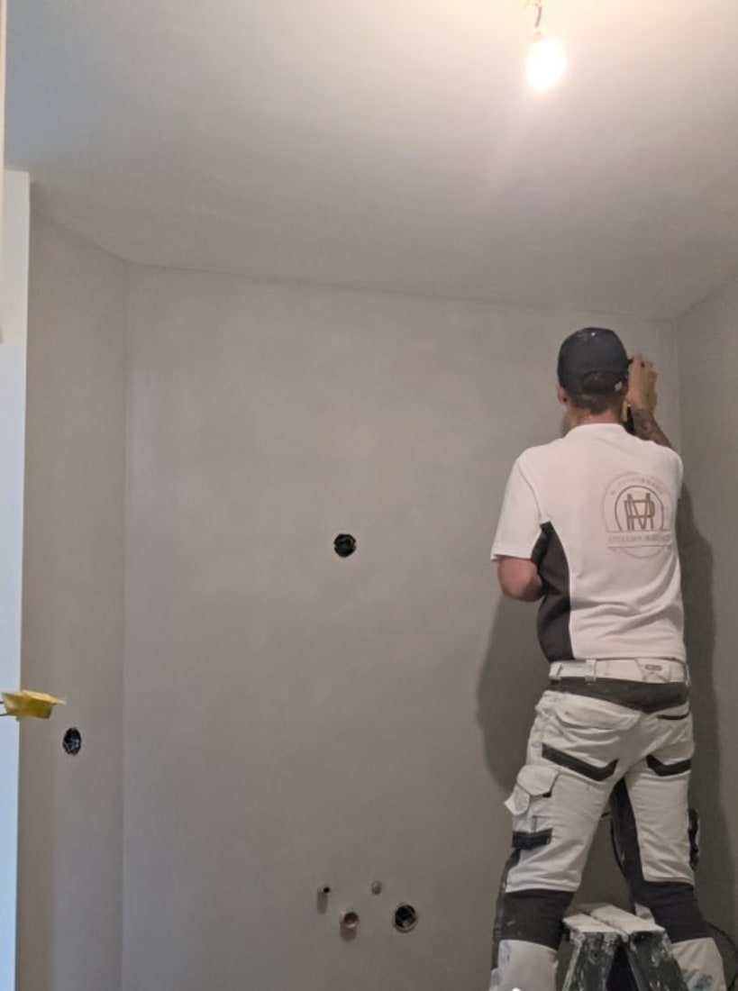 Penninkhof werkt op een trap aan een wand in betonlook, met het logo op de rug van zijn shirt